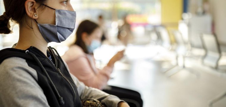 Photo of people sitting in a waiting room, within a healthcare setting. A woman sits in the foreground with a mask over her mouth and nose. The background is blurred but you can see another patient waiting in a mask, and chairs for other patients. 