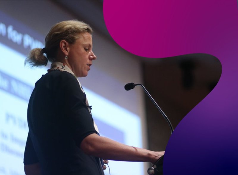 A woman presenting at an event, with PVRI's fluid shapes in a gradient of pink, purple, plum and indigo swirling at the edges. 