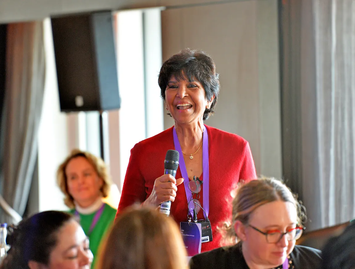 Dr Usha Raj from University of Illinois smiling with a microphone. Taken at PVRI's Women in Pulmonary Hypertension Luncheon, held during the Annual Congress, February 2024. Other women visible in the foreground and background, slightly out of focus.