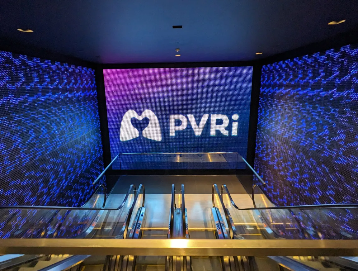 The entrance to the PVRI Annual Conference, with the PVRI logo shown an a giant LED screen at the bottom of a set of escalators. More screens to the sides feature a bright pattern in indigo shades, drawing the eye to the logo. The purple shades are reflected on the shiny floor and ceiling.