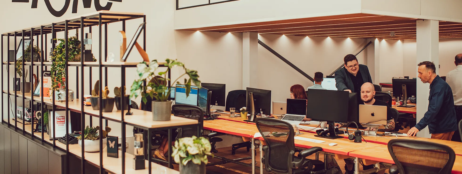 Panoramic shot across the IE studio, Members of the team are seen collaborating. A shelving unit featuring the agency's awards and various plants and other paraphernalia acts as a room divider.